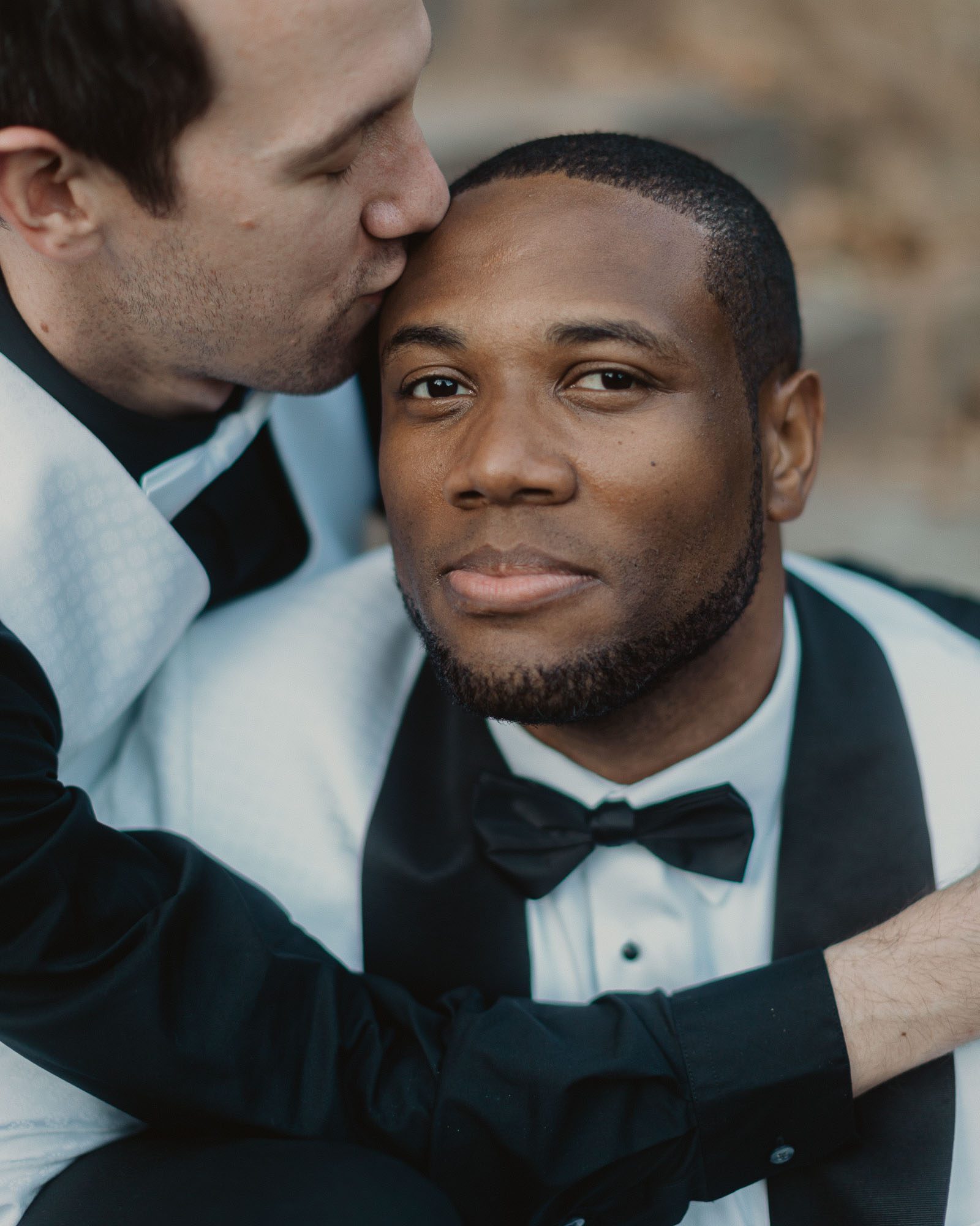 Wedding portraits in historic Atlanta Piedmont Park, LGBTQ male couple in wedding attire embrace while one man kisses the forehead of the other who is looking at the camera.