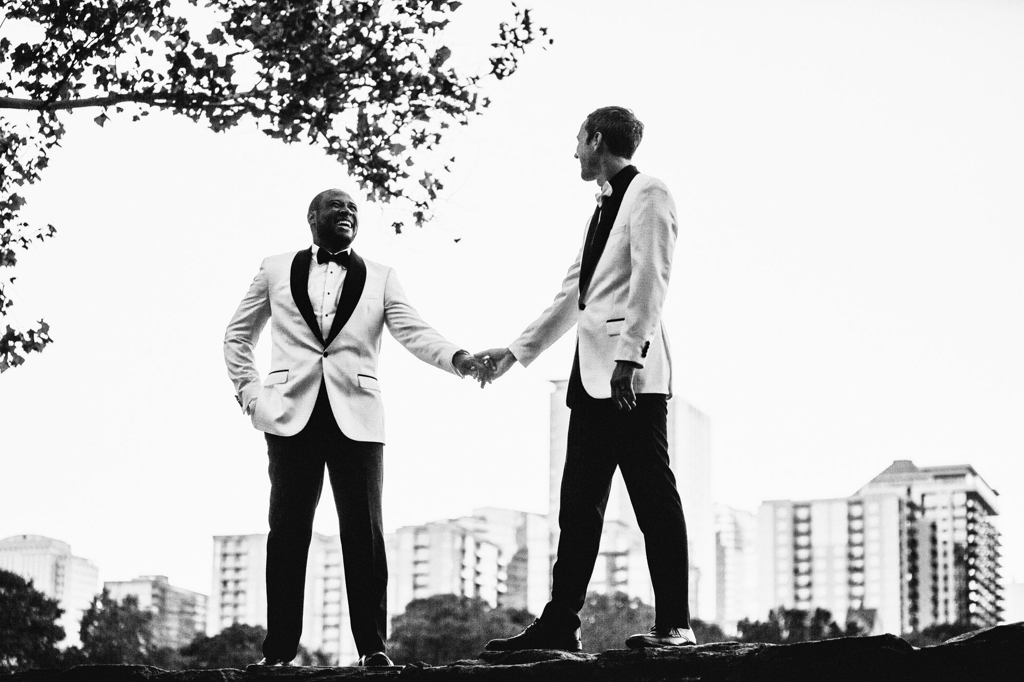 Wedding portraits in historic Atlanta Piedmont Park. LGBTQ male couple in wedding attire standing on a rock wall with Atlanta skyline in background.