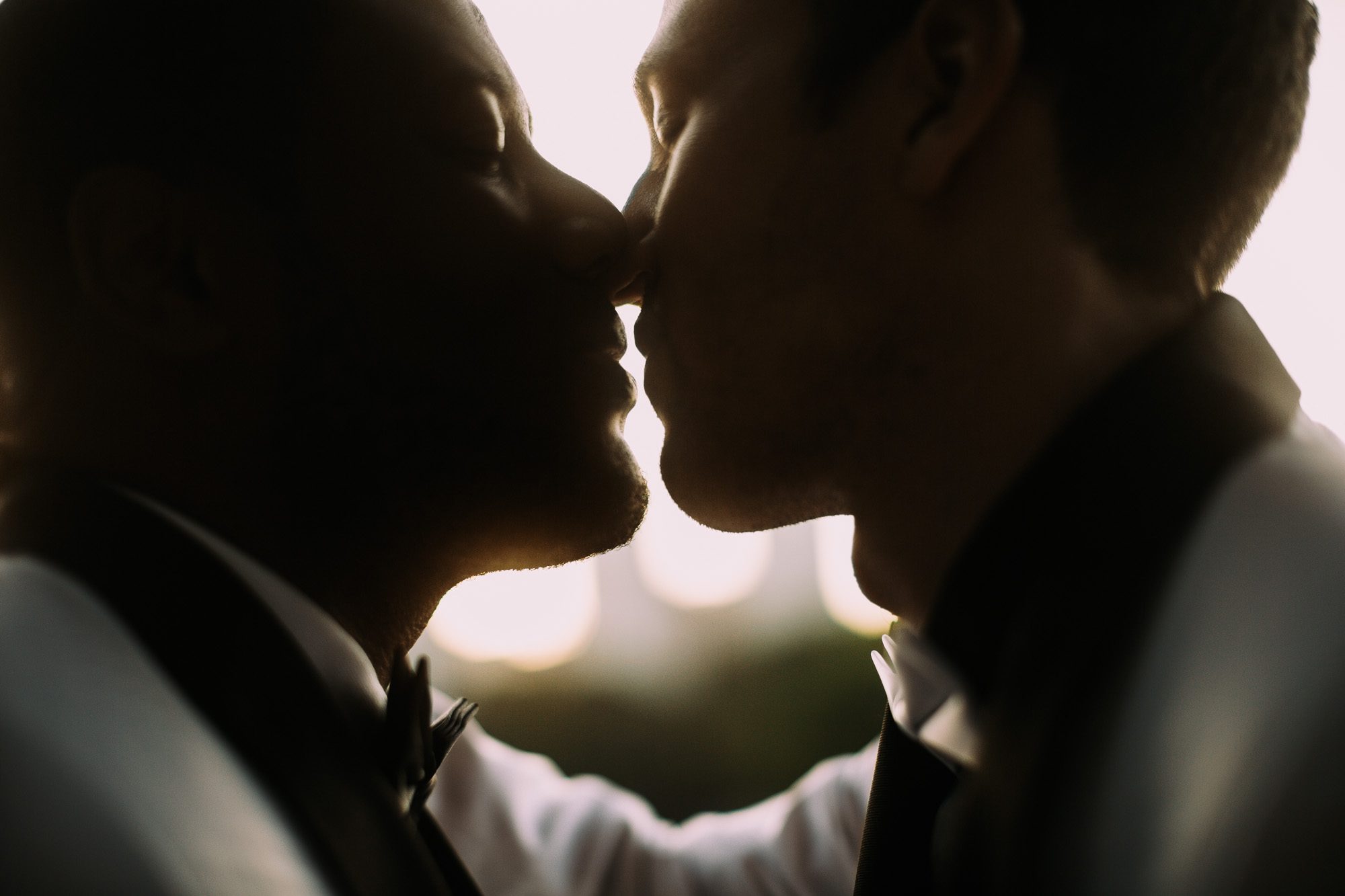 Wedding portraits in historic Atlanta Piedmont Park, LGBTQ male couple in wedding attire are silhouetted against the sky and leaning in for a kiss.