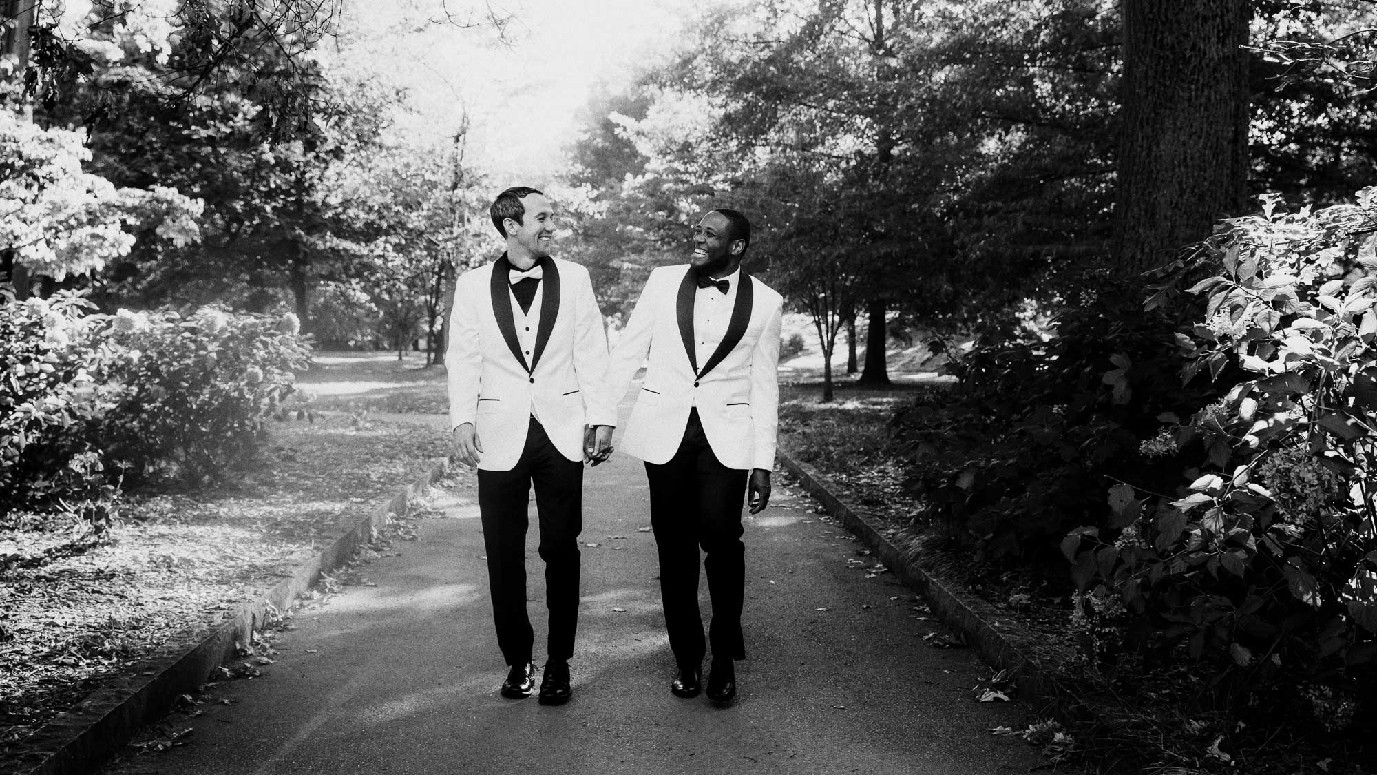 Wedding portraits in historic Atlanta Piedmont Park, LGBTQ male couple in wedding attire hold hands and walk toward the camera while looking at each other and smiling.