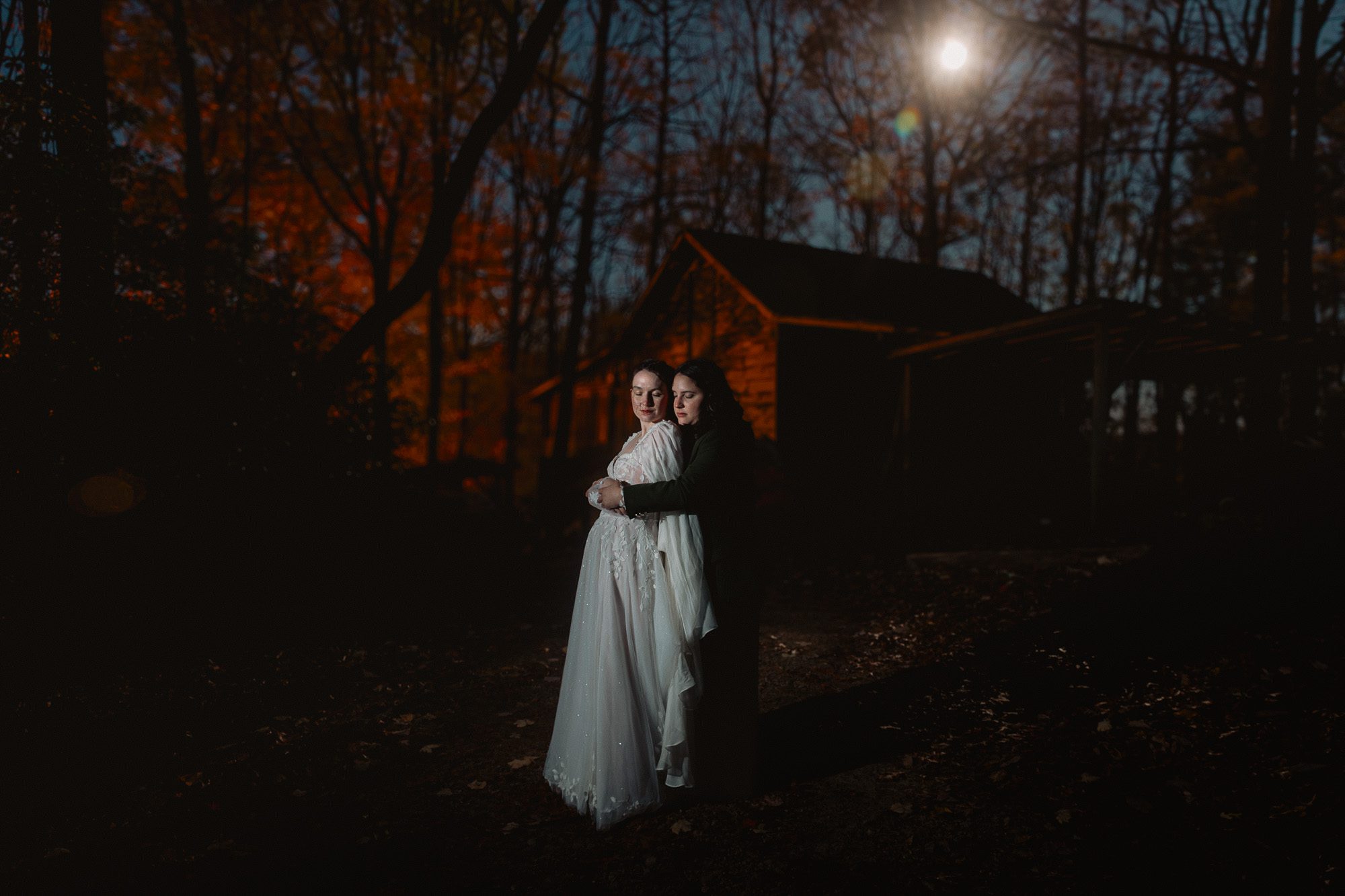 an LGBTQ couple embrace standing outside under the full moon in a wooded area with a barn at the end of a beautiful fall day spent in north Georgia celebrating their elopement