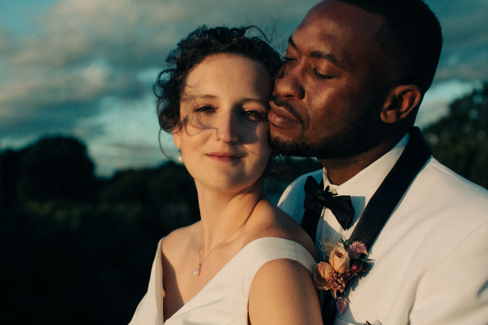 a biracial couple embraces at sunset on their wedding day, the bride is looking at the viewer and the groom is looking at the bride, she is wearing a white wedding gown and he is wearing a white tuxedo and black tie