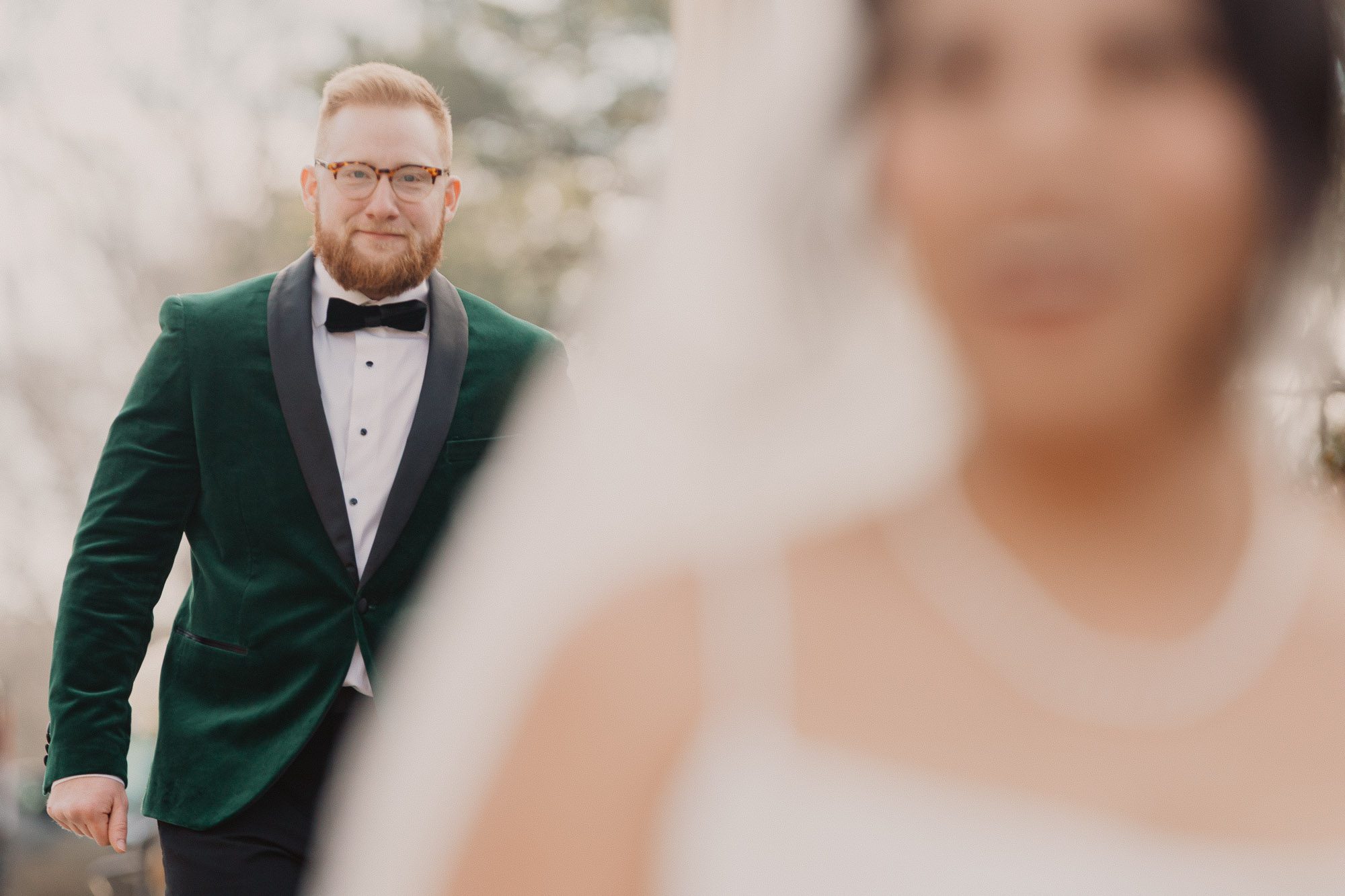 The bride waits with eyes closed as the groom approaches from behind in soft light.