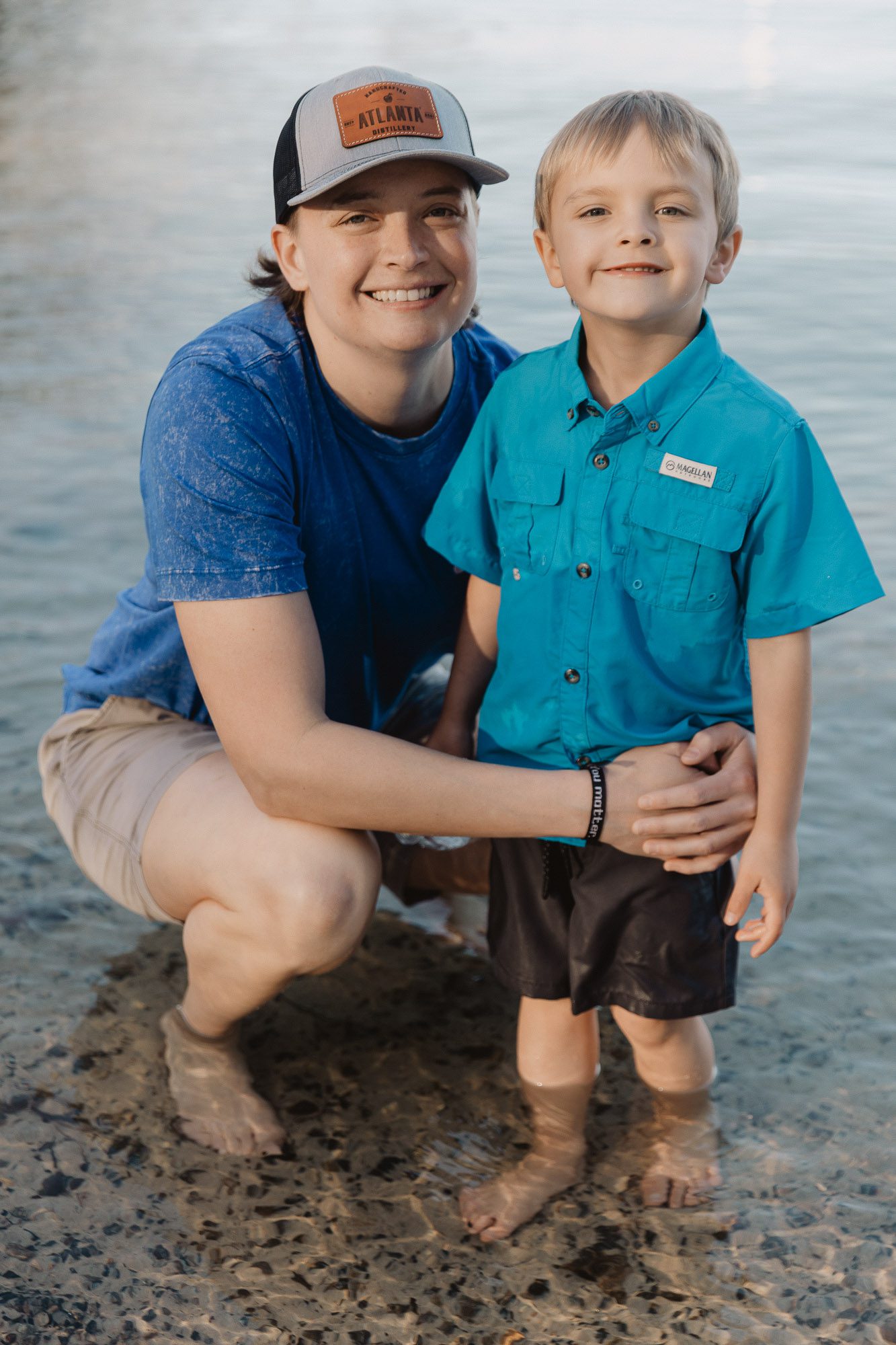 a mother kneels in the water and embraces her small child on the beach at Lake Lanier during their lifestyle family photo session
