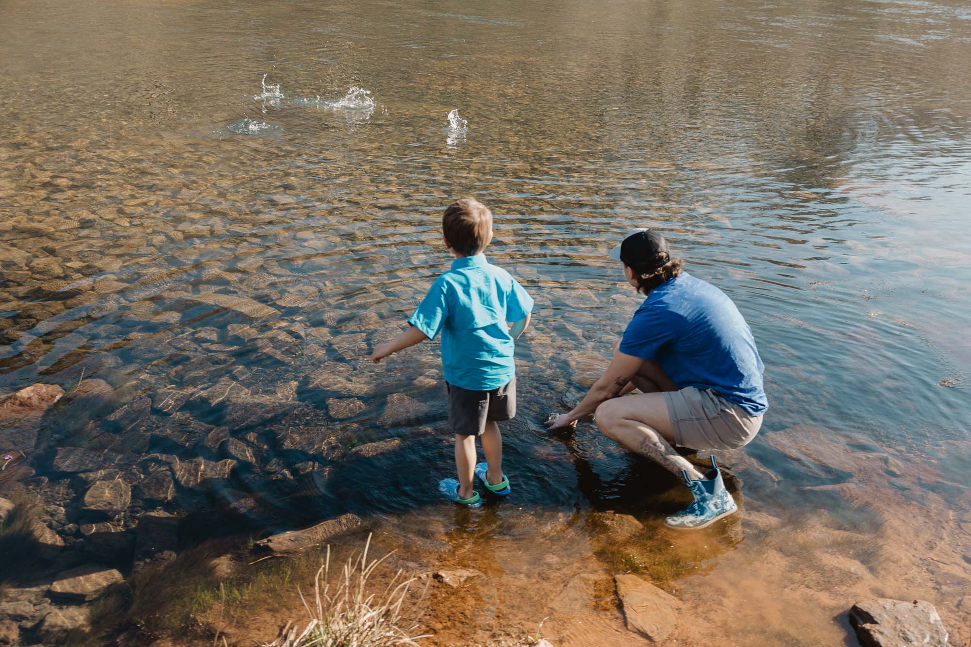 a small child throws a rock into the Chattahoochee river while his mother kneels and watches during their active lifestyle family photo session