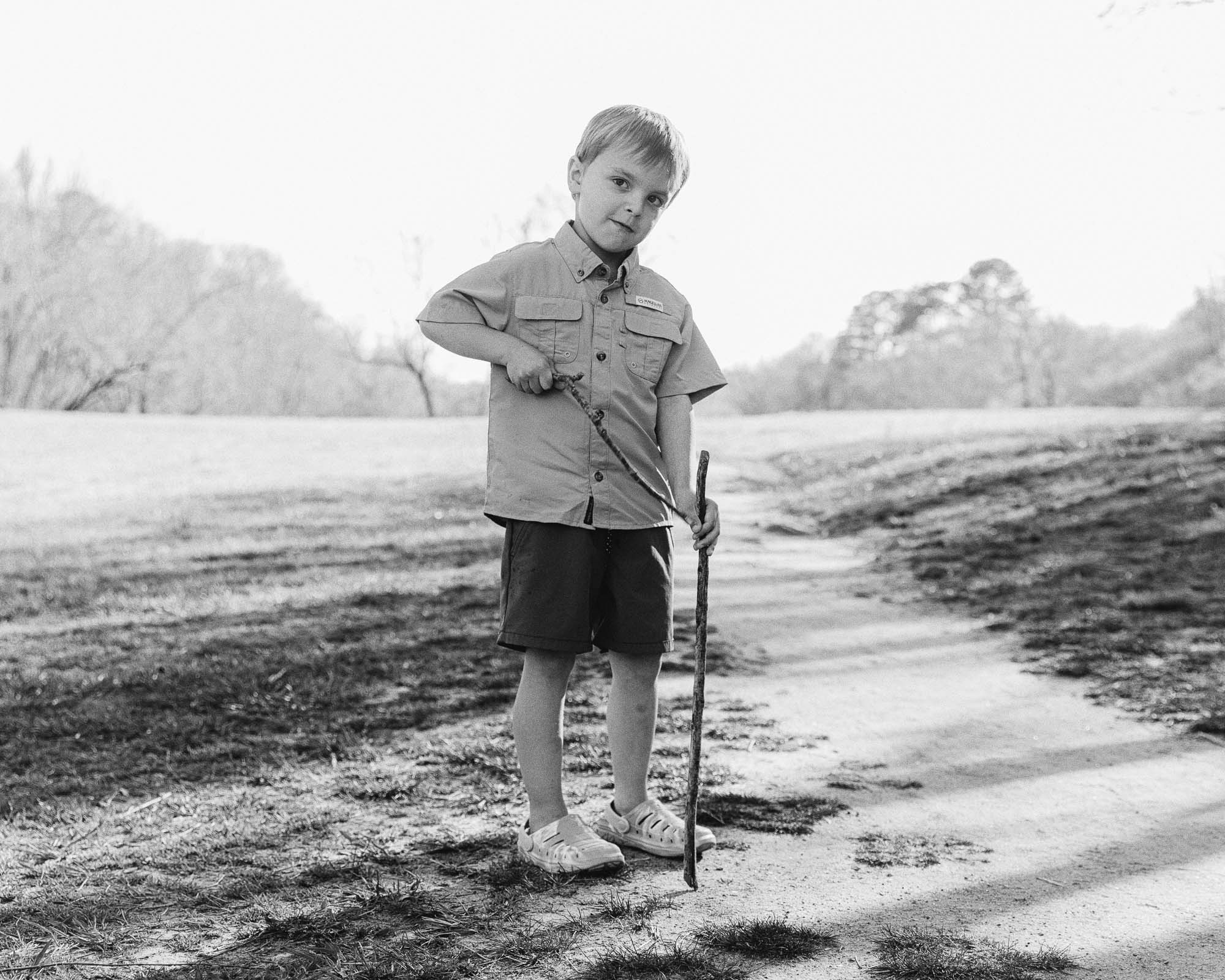 a small child stands and poses holding a stick he found at the park during a lifestyle family photo session