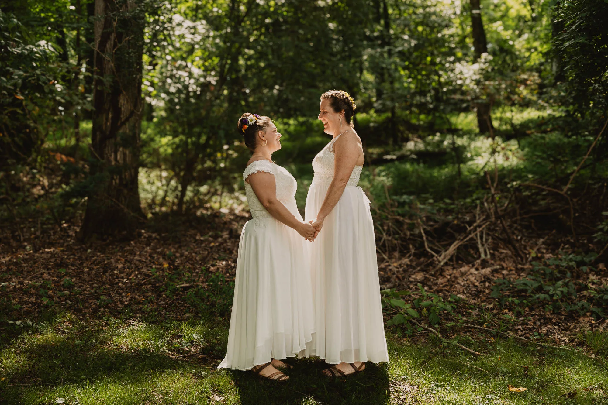 two brides wearing white dresses hold hands while standing in the sunlight near the forest at Joe Wheeler State Park in Alabama