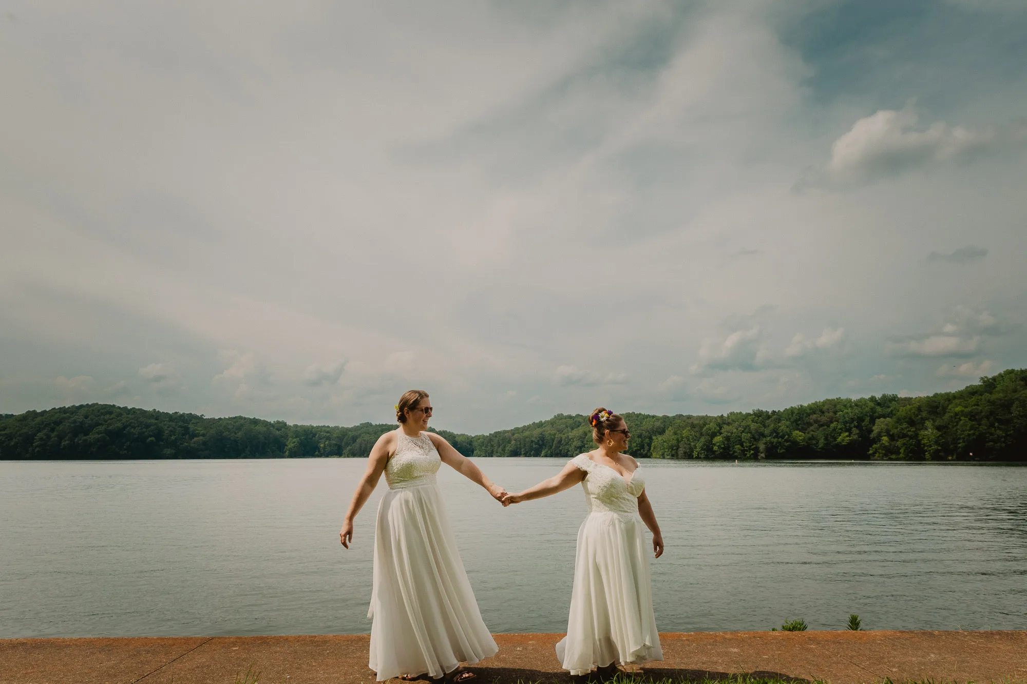 two brides wearing white dresses hold hands and walk along the shoreline of Wheeler Lake at Joe Wheeler State Park in northwest Alabama