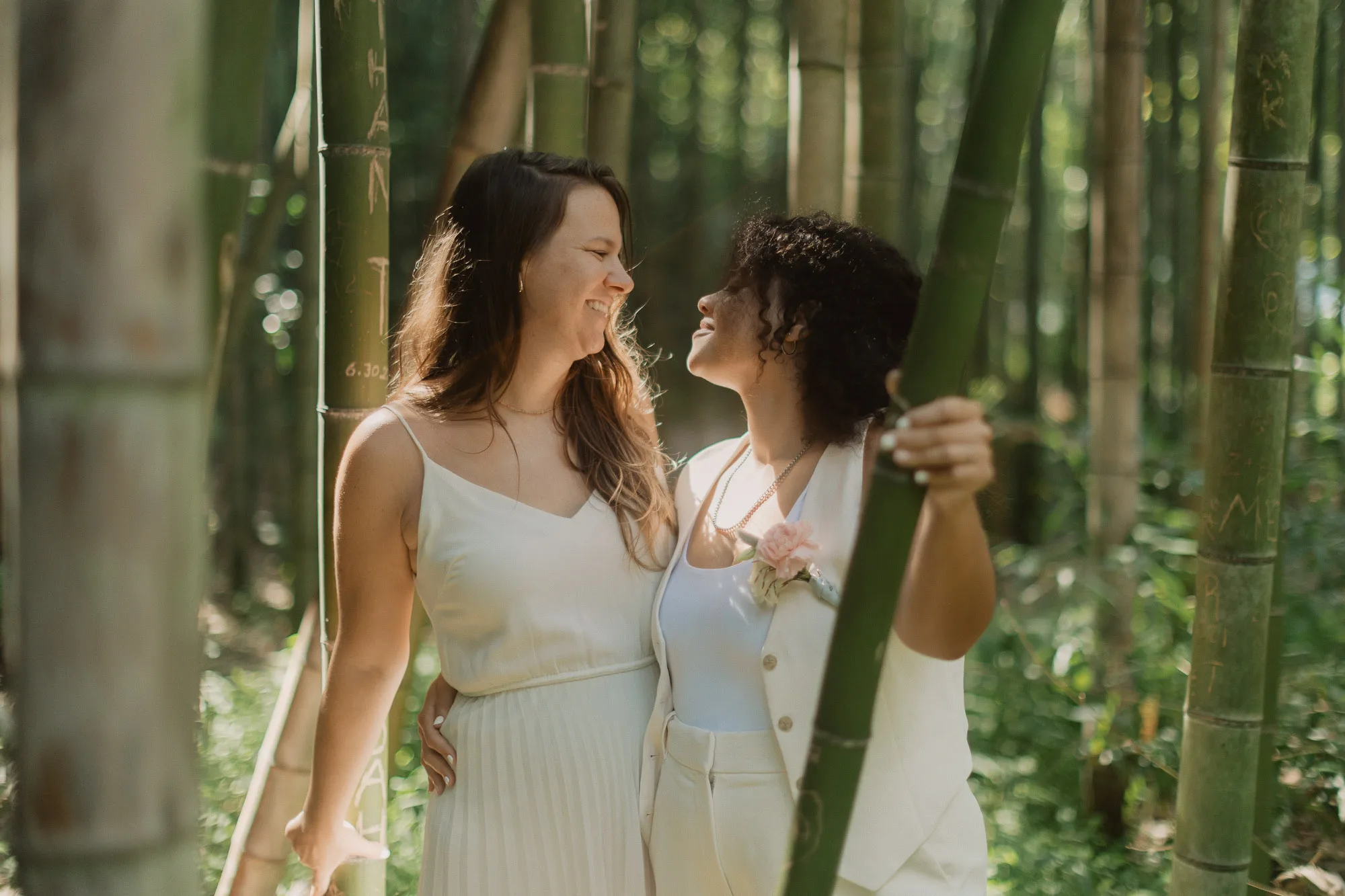 two brides wearing white stand close and look at each other on their wedding day in the bamboo forest on the chattahoochee river in atlanta