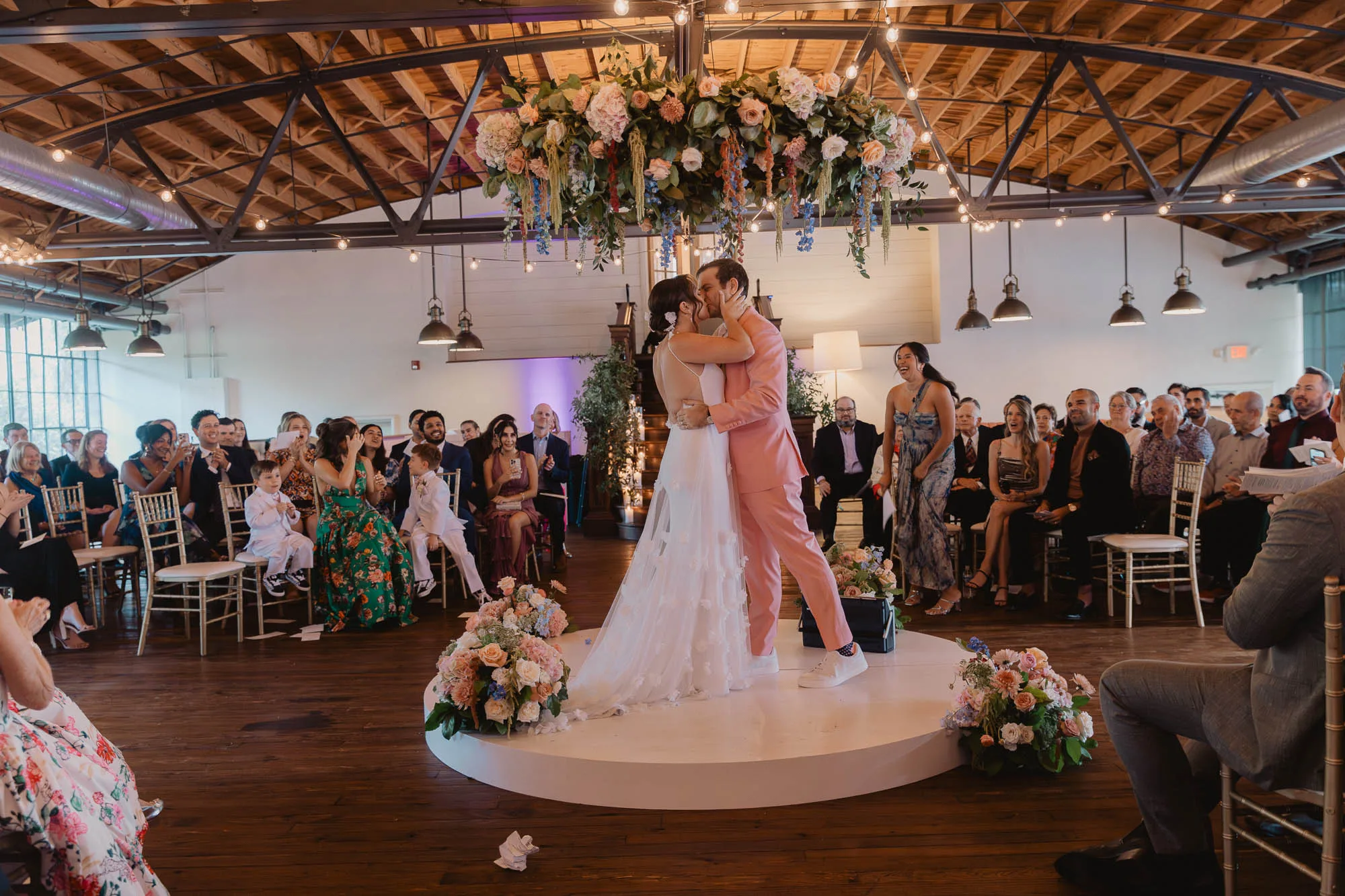 a bride wearing a white dress and a groom wearing a pink tuxedo share their first kiss during their wedding ceremony at Summerour Studio in Atlanta