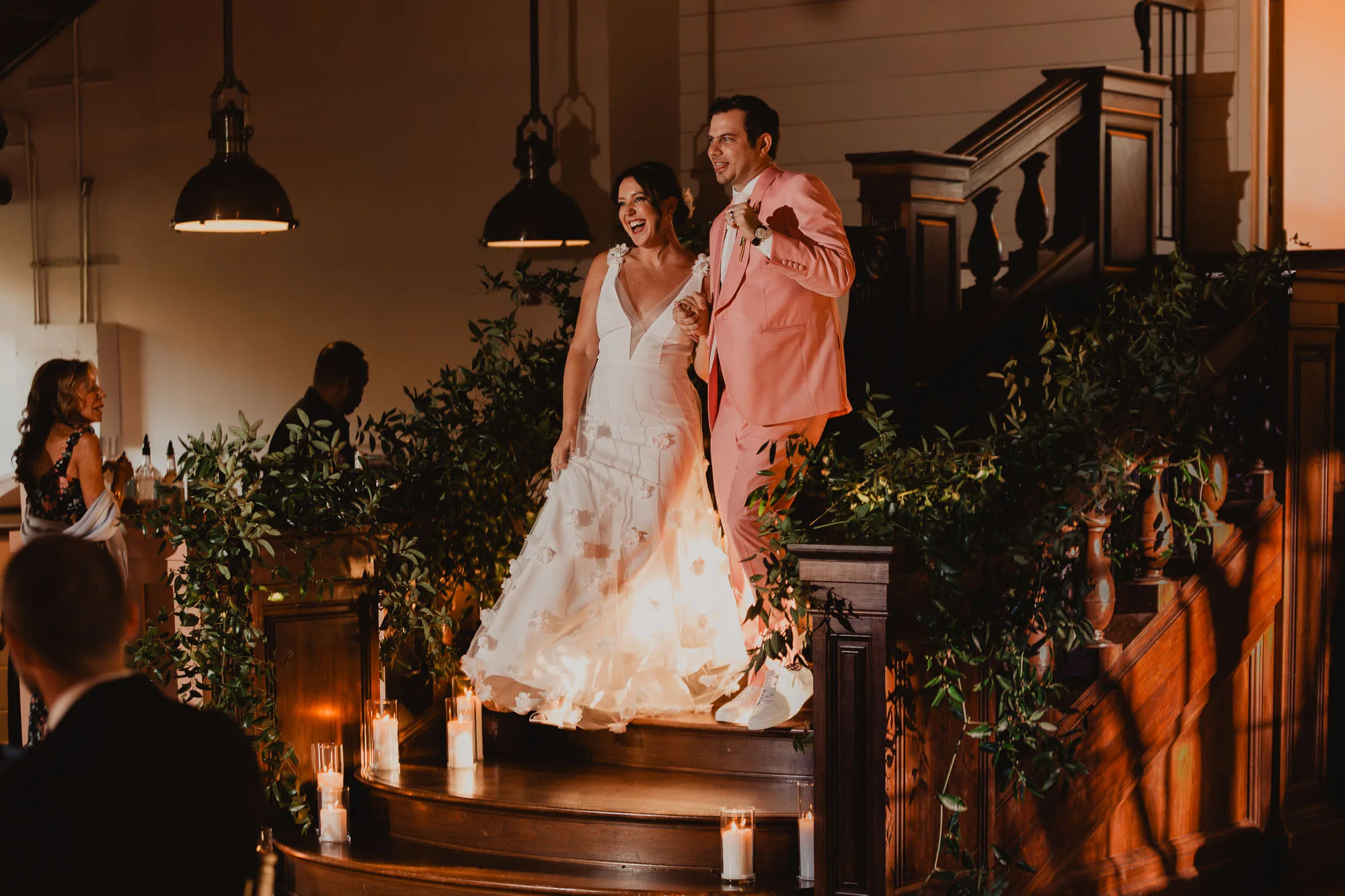 a bride wearing a white dress and a groom wearing a pink tuxedo make their grand entrance coming down the grand staircase at their wedding reception at Summerour Studio in Atlanta