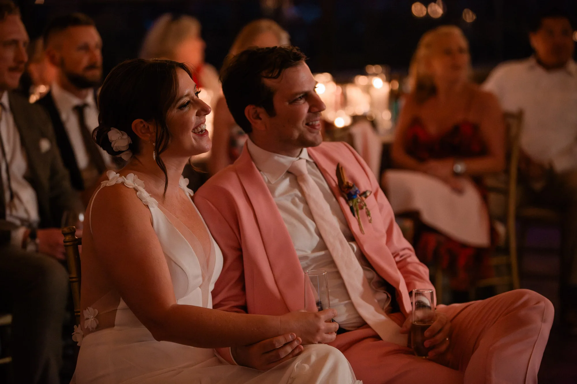a bride wearing a white dress and a groom wearing a pink tuxedo are seated and listening to toasts at their wedding ceremony at Summerour Studio in Atlanta
