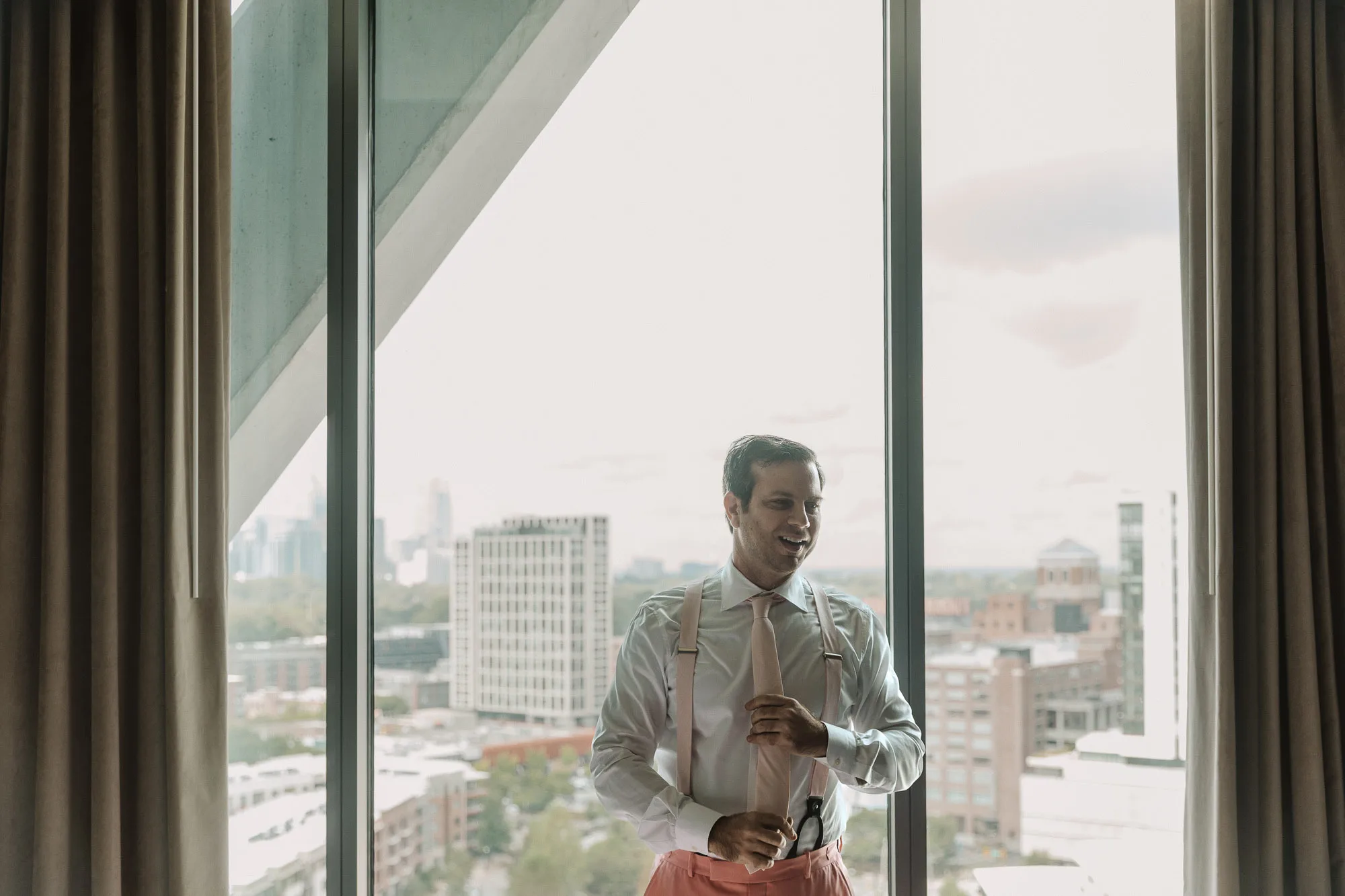 an Atlanta groom adjusts his tie while standing in front of a large window overlooking the skyline at the Forth Hotel