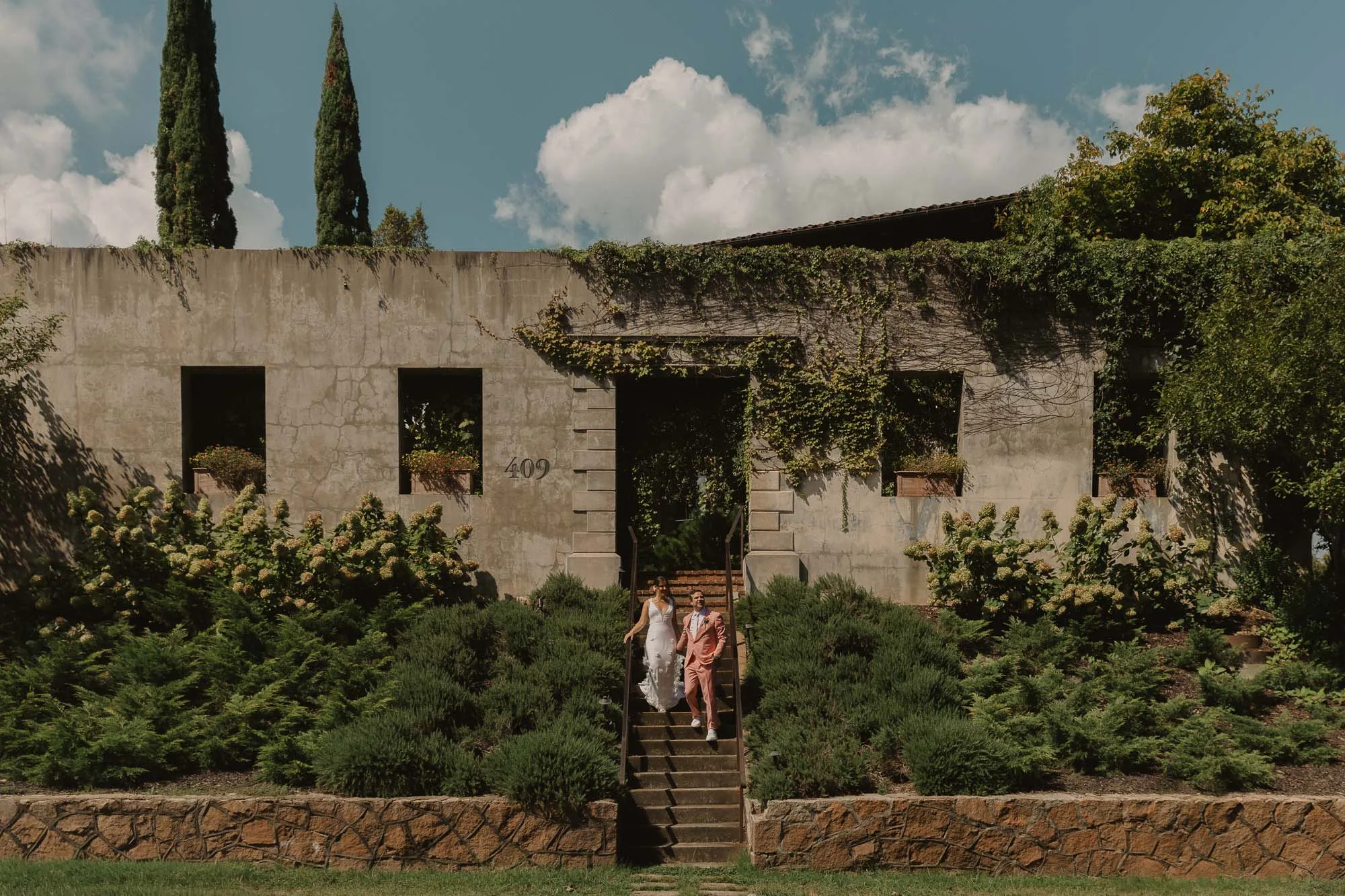 a bride wearing a white dress and a groom wearing a pink tuxedo walk down the stairs holding hands in front of Summerour Studio in Atlanta