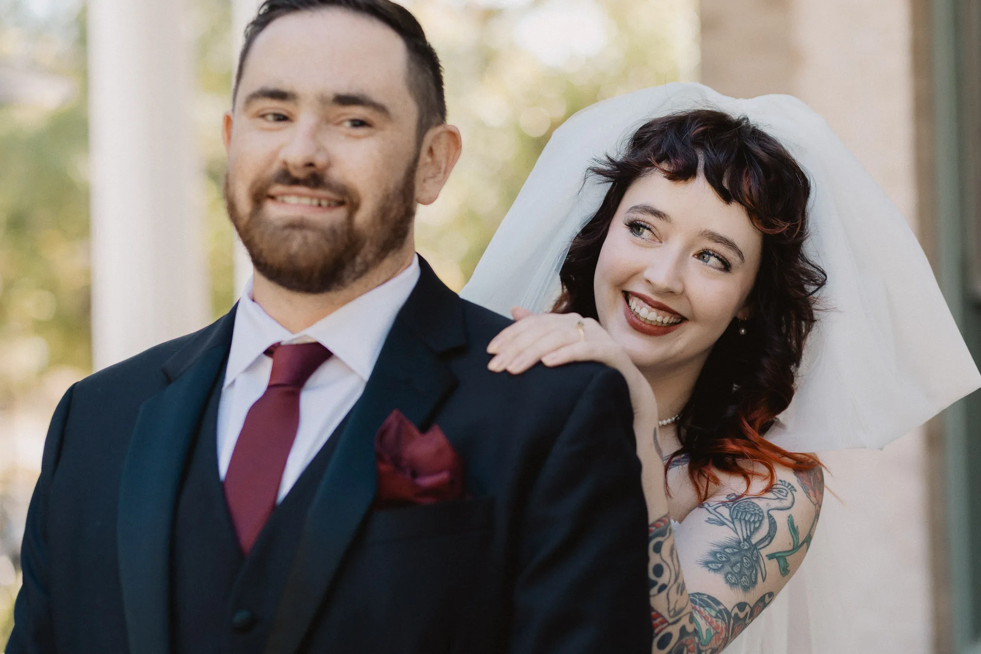 a smiling bride wearing a white veil peeks over the shoulder of a groom wearing a black tuxedo and burgundy tie on the porch of The Wrecking Bar in Atlanta